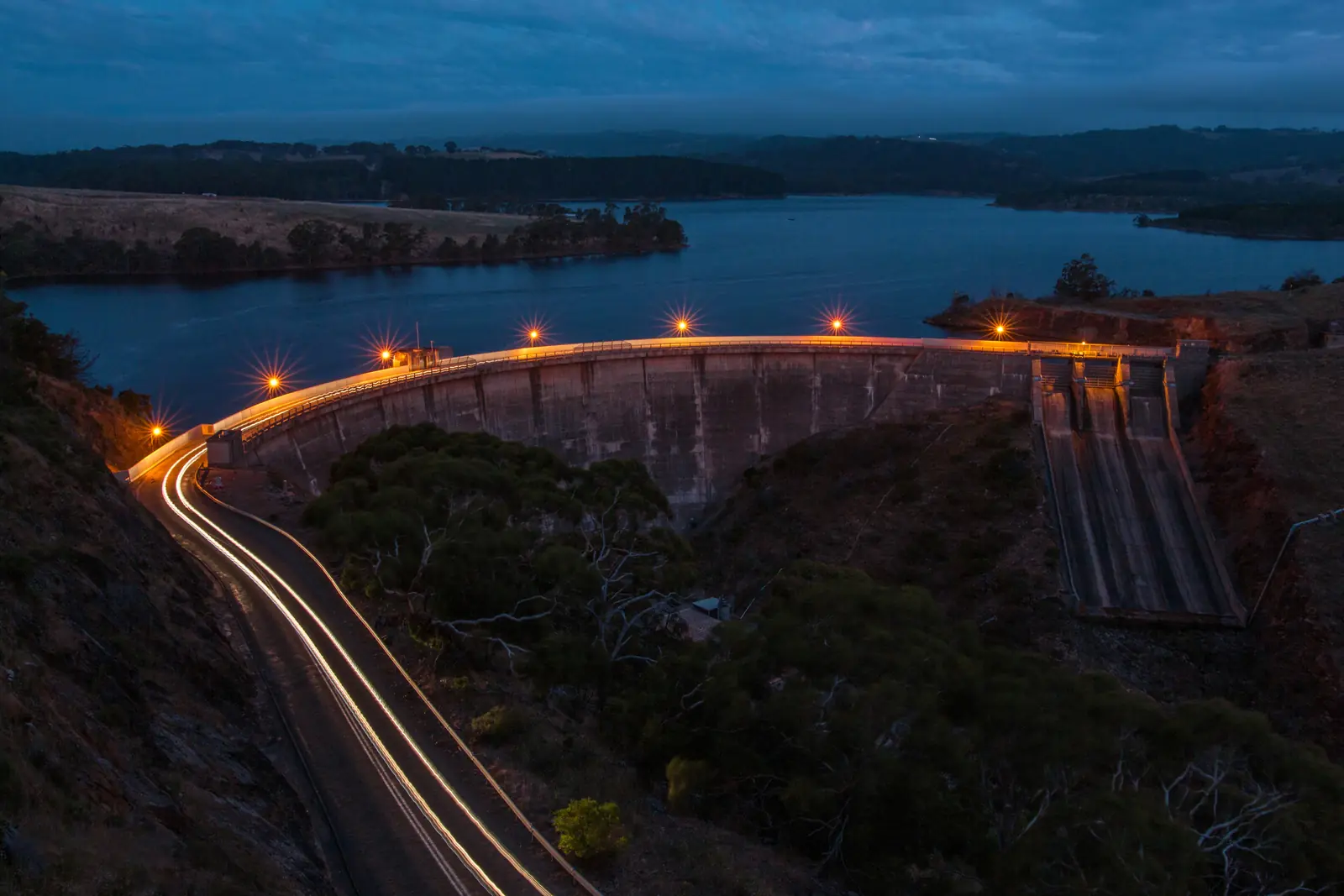 Myponga Reservoir dam wall at dusk, car light trails on the road beside it